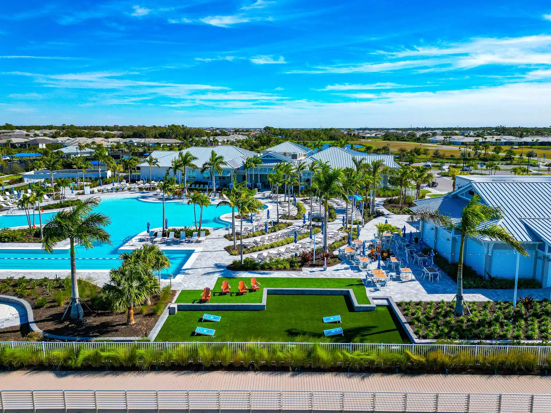 The BeachWalk clubhouse with its white standing-seam metal roofs, seen across the resort pool and lap pool, with rows of loungers and blue umbrellas, a dining terrace with orange chairs, and a lawn set with cornhole boards and orange Adirondack chairs.
