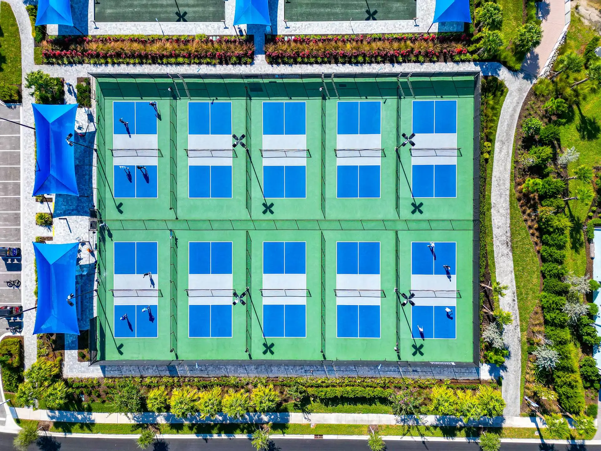 Overhead view of ten pickleball courts at BeachWalk, blue playing surfaces on green surrounds in two rows of five, with players mid-game, blue shade sails along one side, and bocce and tennis courts beyond.