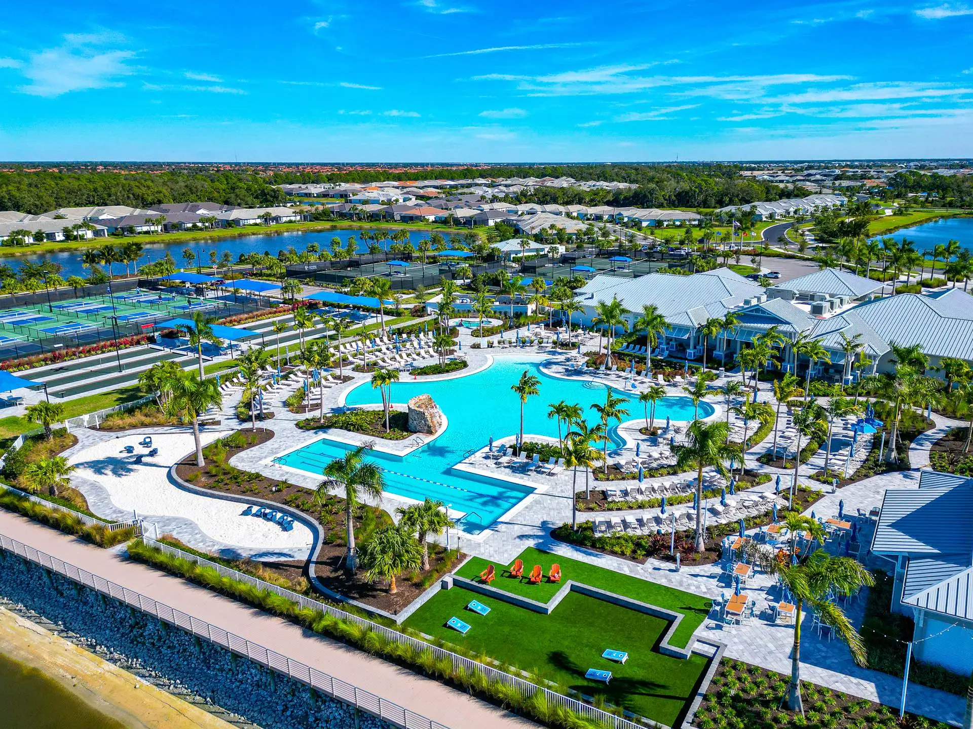 The BeachWalk pool deck from the air: the freeform resort pool and a separate lap pool with swim lanes, a white sand beach edge with loungers, racquet courts and bocce lanes to one side, and the clubhouse and dining terrace beyond.