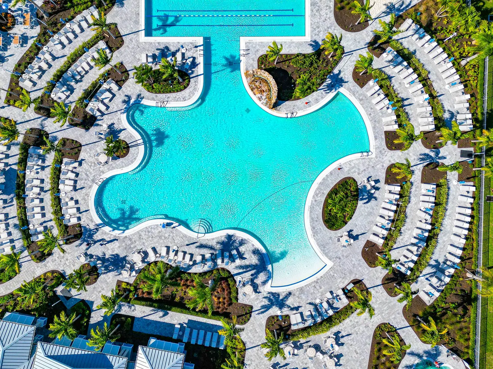 Overhead view of the BeachWalk resort pool: a freeform turquoise pool with a separate lap-lane section and a rock waterfall, planted palm islands, and concentric arcs of white loungers on grey paver decking.