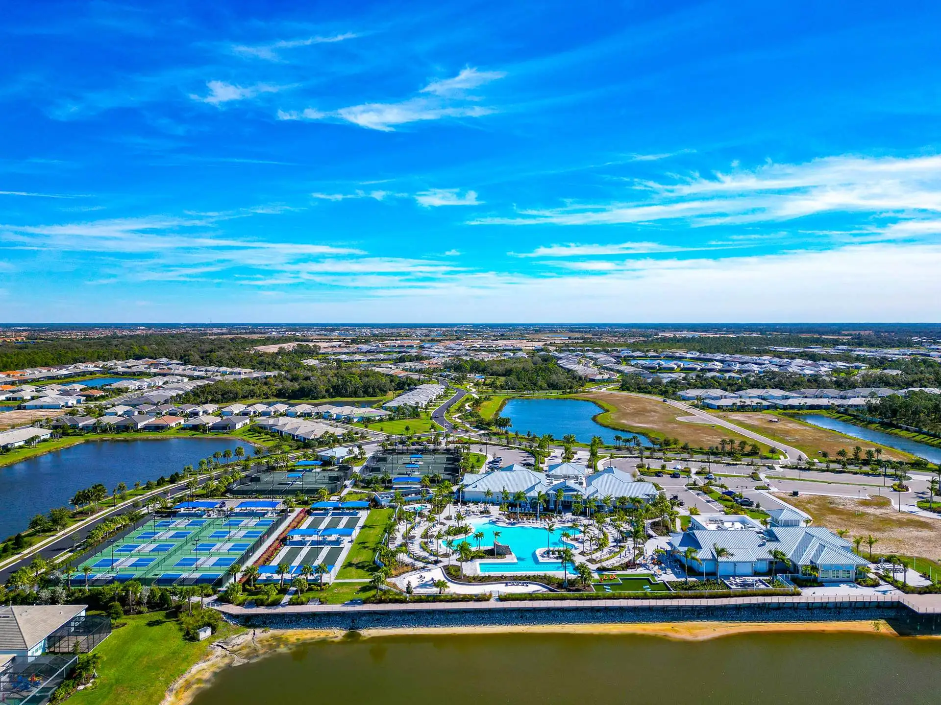 Aerial view over the BeachWalk amenity campus: a resort pool ringed with palms, pickleball and tennis courts under blue shade sails, bocce lanes, a white metal-roofed clubhouse, and a lake and boardwalk in the foreground with single-story homes stretching beyond.