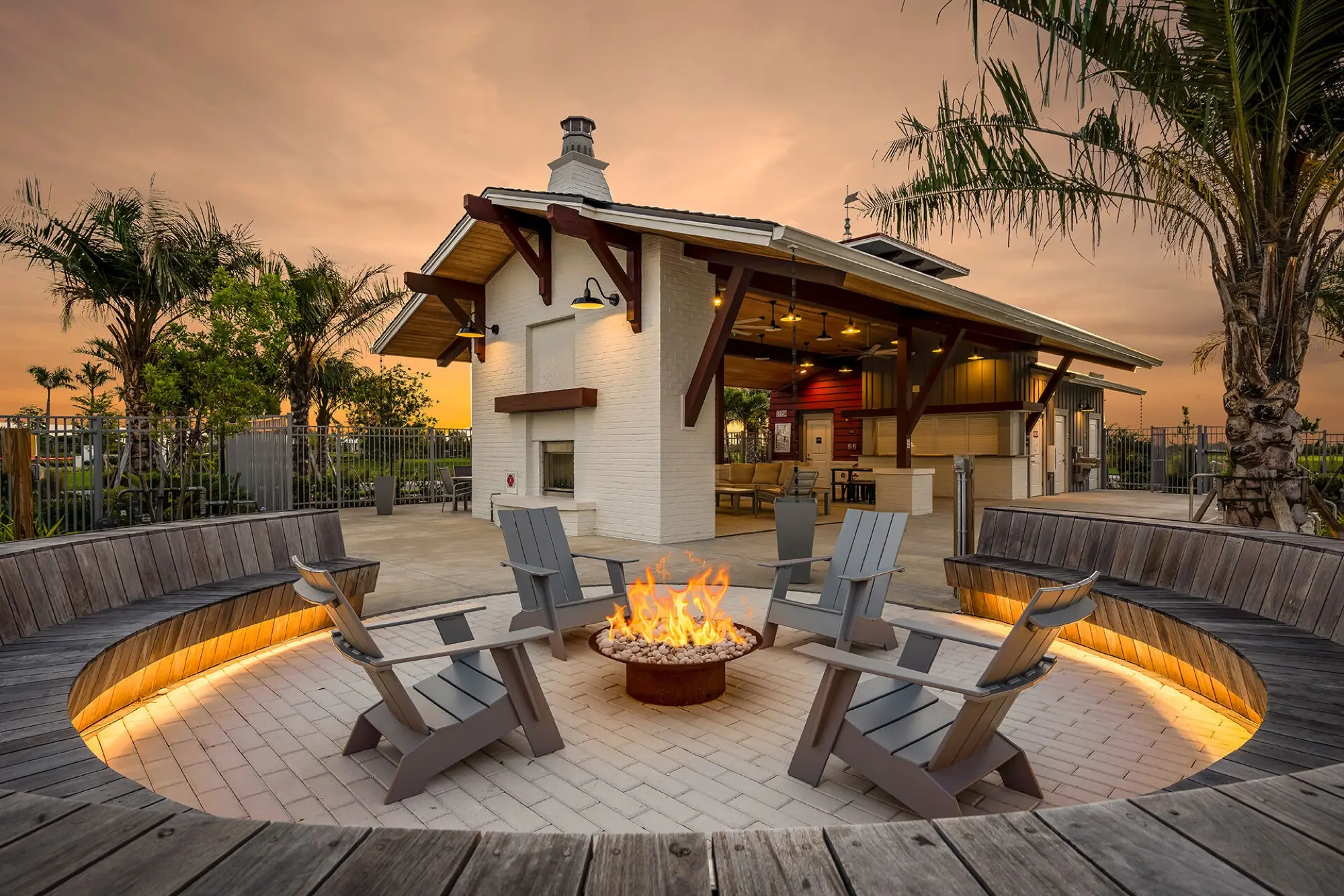 A sunken firepit lounge at Lakespur at sunset, ringed by a curved wooden bench and Adirondack chairs in front of the covered pavilion