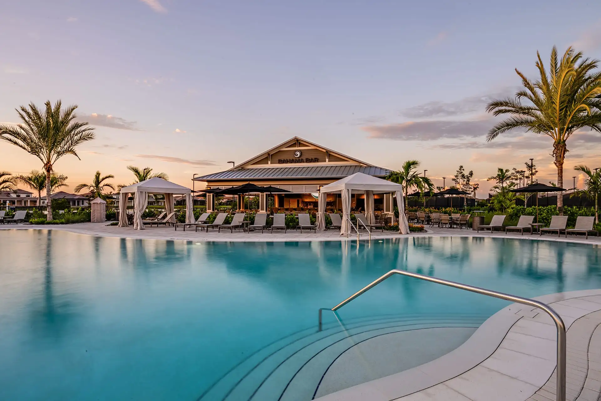 The resort pool at Esplanade at dusk, with the gabled Bahama Bar pavilion lit beyond it, white cabanas and rows of loungers, and palms against a violet and gold sky